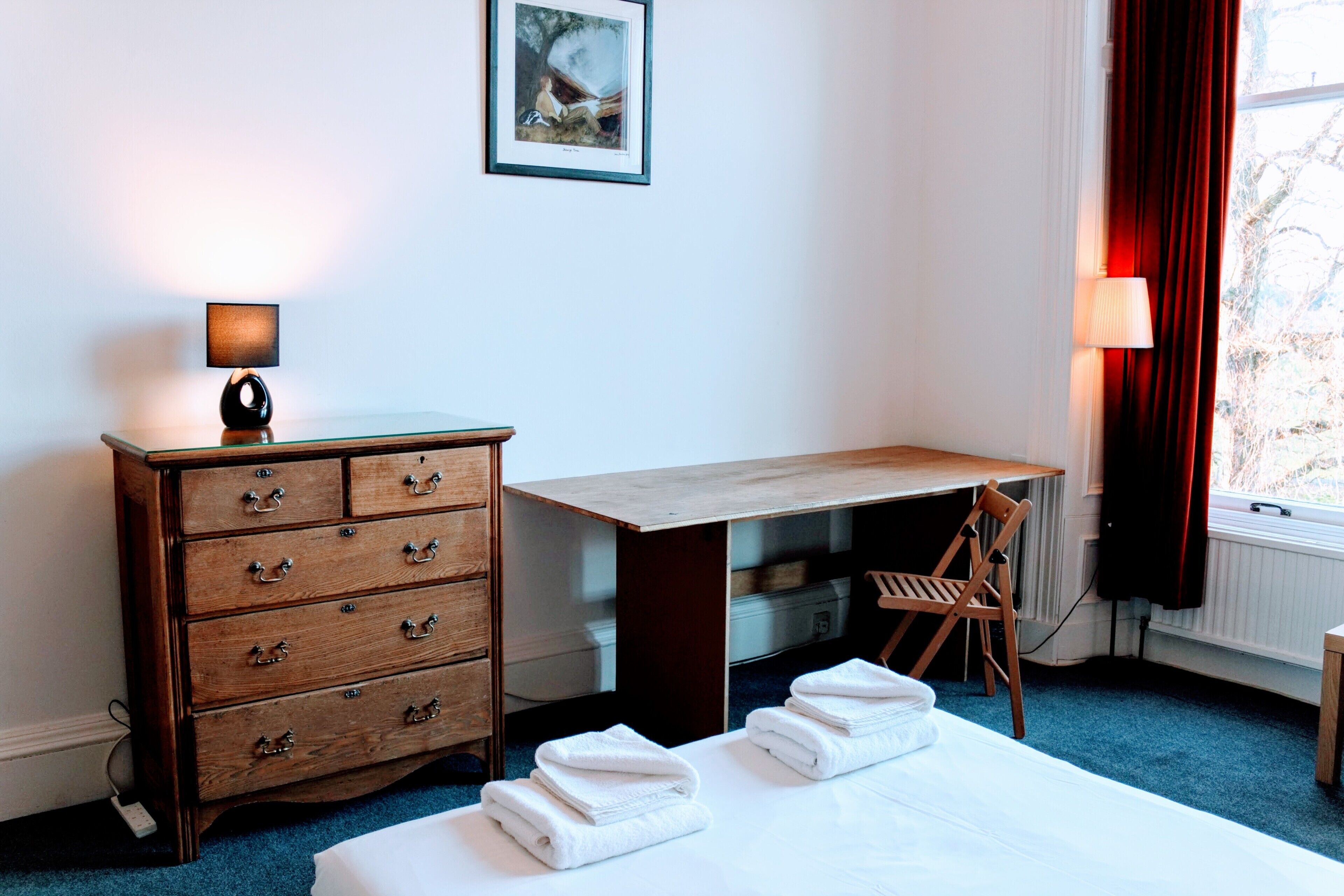 Quarto de hotel com penteadeira de madeira, escrivaninha, cadeira, cama com toalhas, luminária, quadro na parede e janela com cortinas.