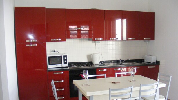 A compact kitchen with red cabinets, a white countertop, a microwave, a kettle, and a dining table with four chairs.