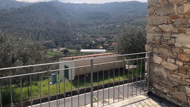Un balcón con muro de piedra, baranda metálica y vista a un valle con casas y árboles.