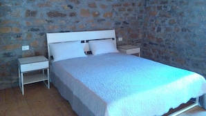 Un dormitorio con pared de piedra, una cama blanca y una mesita de noche con lámpara.