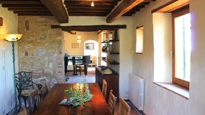 Un comedor rústico con una mesa de madera, sillas y una pared de piedra. Se ve un piano al fondo y una ventana a la derecha.