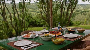 Mesa de comedor al aire libre preparada con platos, fuentes y cubiertos, rodeada de sillas de mimbre, con una vista panorámica de vegetación y colinas al fondo.