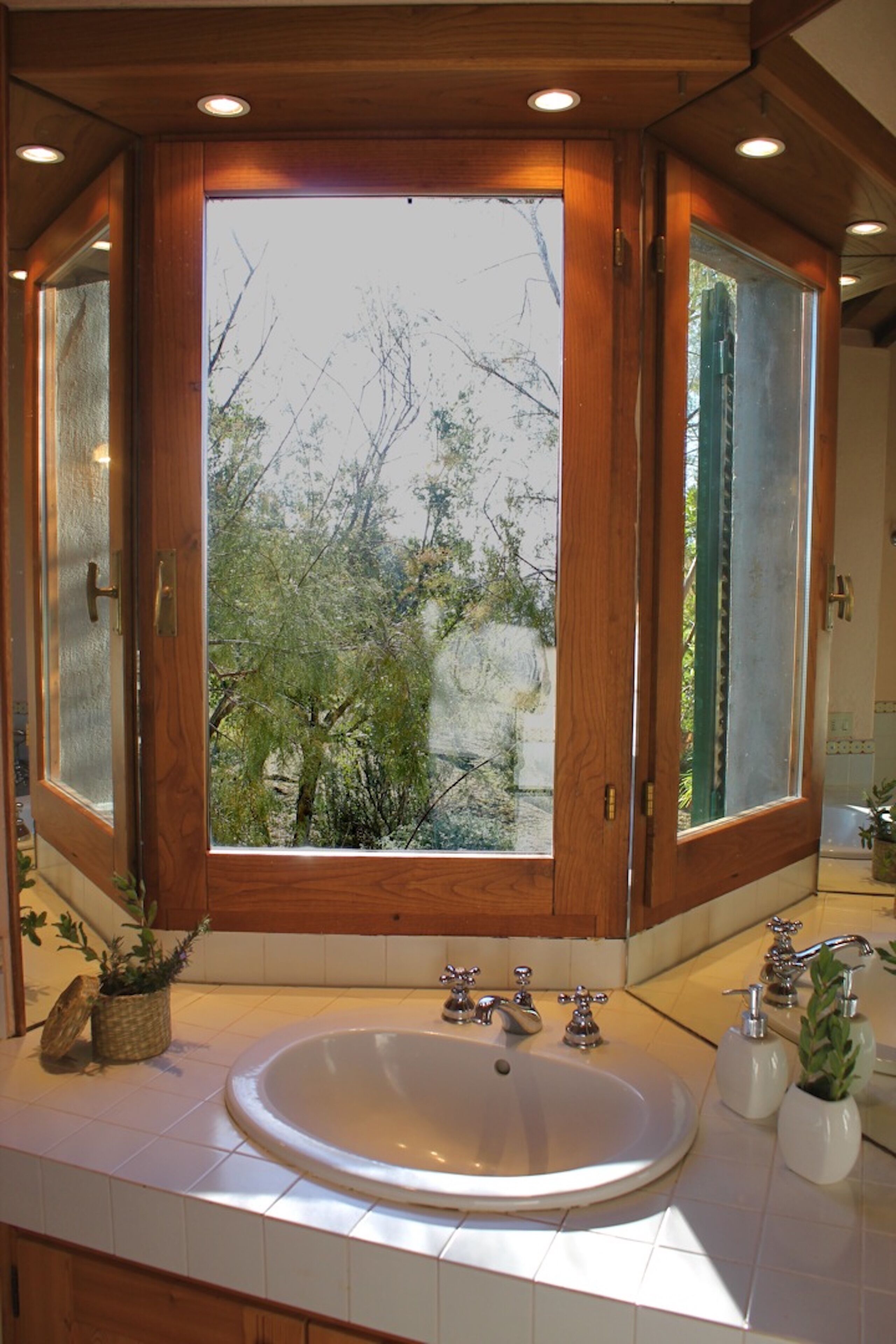 A bathroom with a large window, wooden cabinetry, and a sink with two faucets.