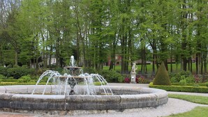 Une fontaine circulaire en pierre, avec des jets d’eau, se trouve dans un jardin aménagé, agrémenté d’arbres et d’une statue en arrière-plan.