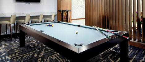 A pool table with a billiard cue, a television mounted on the wall, and a seating area with chairs.