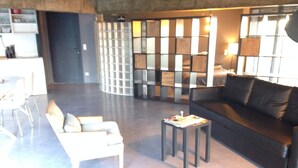 A modern living room with a black sofa, white armchairs, and a small table with a magazine. A glass partition separates the living area from a kitchen area in the background. - Loft on Mont St Clair with private pool and sea view - SETE (Sète)