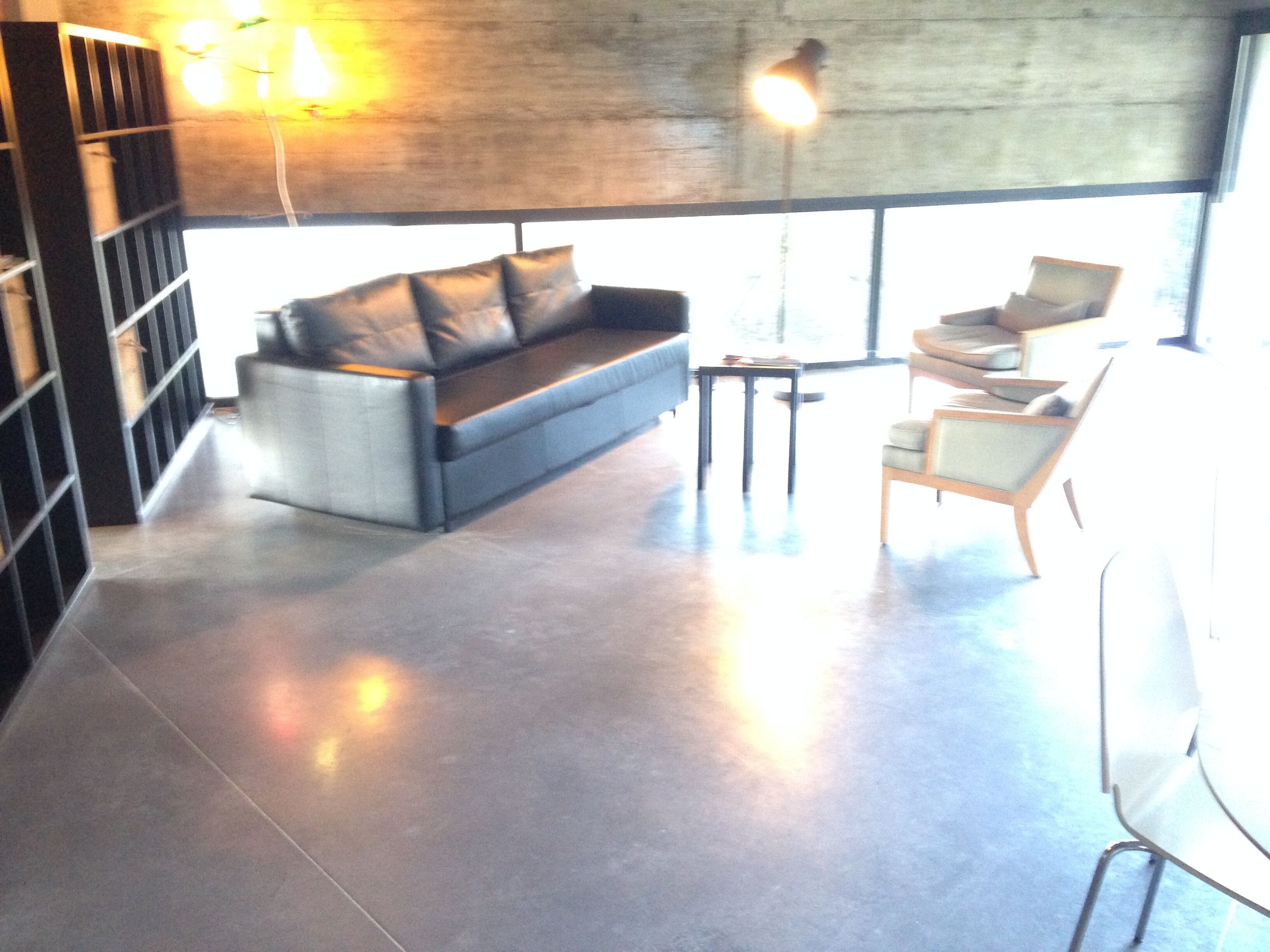 Un salon moderne avec un canapé, un fauteuil et une petite table. Une bibliothèque se trouve à gauche et une grande fenêtre à droite.
