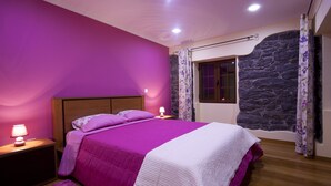 A bedroom with a purple wall, a stone accent wall, a wooden headboard, a bed with a purple and white duvet, a nightstand with a lamp, and a window with a curtain. - Casa do Barco, Rustic house with salt pool and sea view (Arco da Calheta)