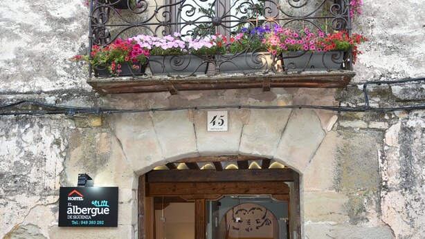 A hostel entrance with a stone facade, a balcony with wrought iron railing, and a sign indicating the hostel name and contact number.