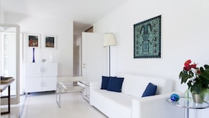 Una sala de estar moderna con un sofá blanco, una mesa de centro de vidrio y una lámpara de pie. Hay un mueble blanco con un jarrón azul y un marco de fotos encima. Un cuadro adorna la pared, y una planta en maceta con flores rojas se encuentra a la derecha.