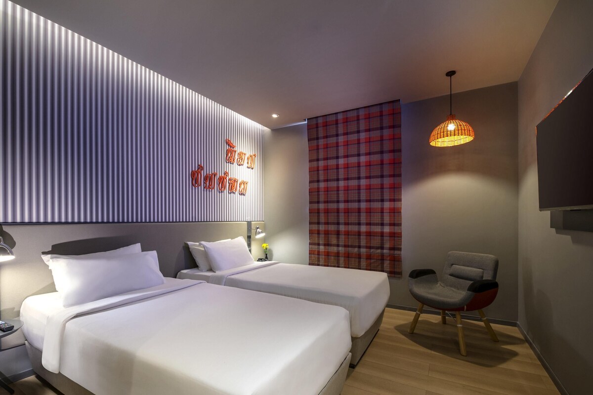 Een moderne hotelkamer met twee bedden, een rood geruit gordijn, een hanglamp en een flatscreen televisie die aan de muur is bevestigd.