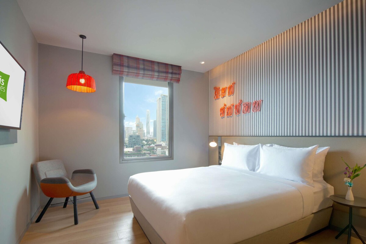 Een moderne hotelkamer met een groot bed, stadszicht, een rode hanglamp en een geruit gordijn.