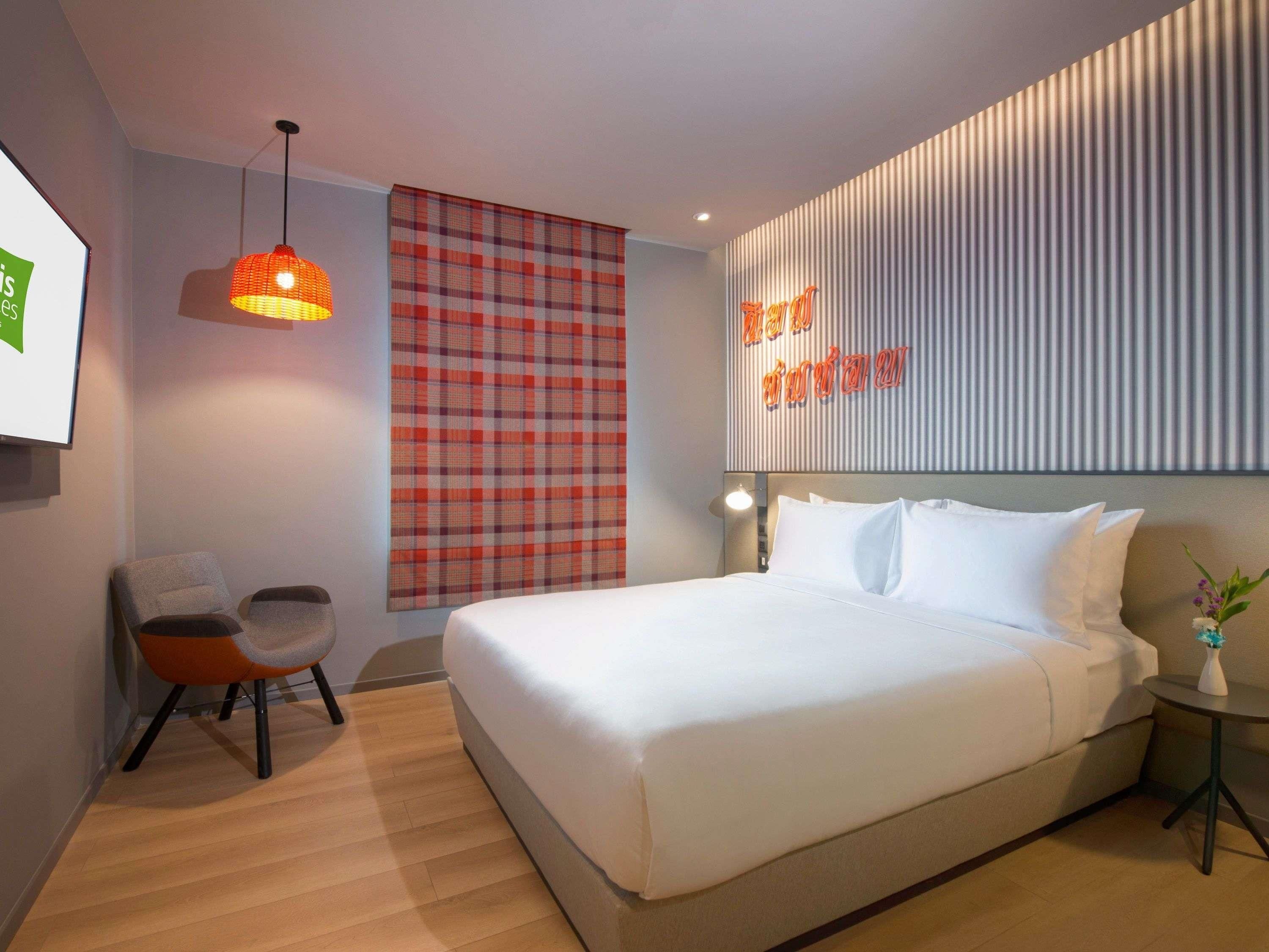 A modern hotel room with a large bed, a seating area with two chairs, a wall-mounted TV, and a decorative wall panel.