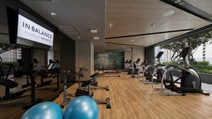 Egy modern, edzőgépekkel felszerelt fitneszterem, ahol egy nagy képernyőn a „NOVOTEL IN BALANCE FITNESS” felirat látható, és nagy ablakokból a városra nyílik kilátás.