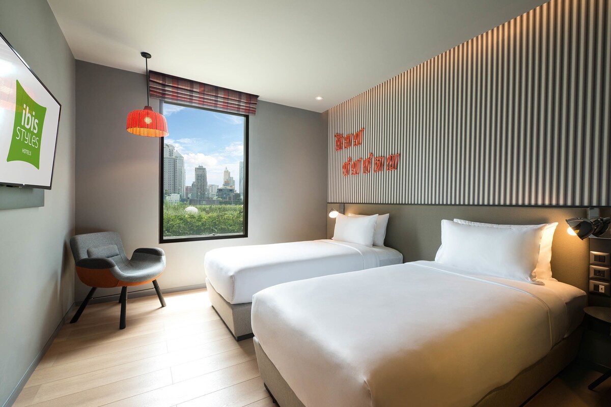 A modern hotel room with two beds, a chair, a city view, and a wall-mounted light fixture.