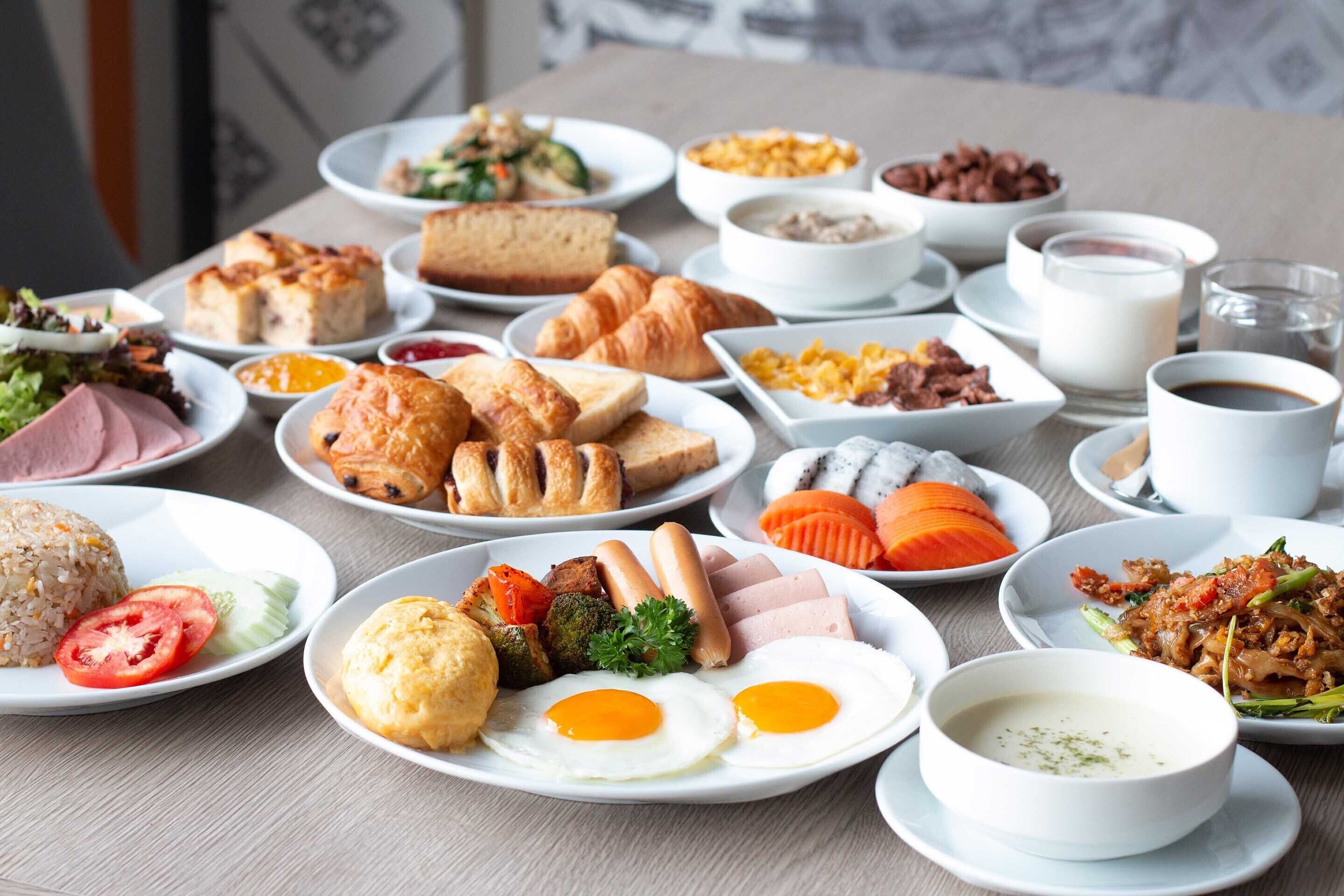 Une variété de plats pour le petit-déjeuner, notamment des œufs, du jambon, des croissants et des viennoiseries, sont disposés sur une table.