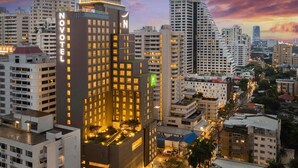 Egy modern hotel, melynek feltűnő „NOVOTEL” felirata díszíti, és amelyet magas épületek vesznek körül, a háttérben pedig a naplemente égboltja látható.