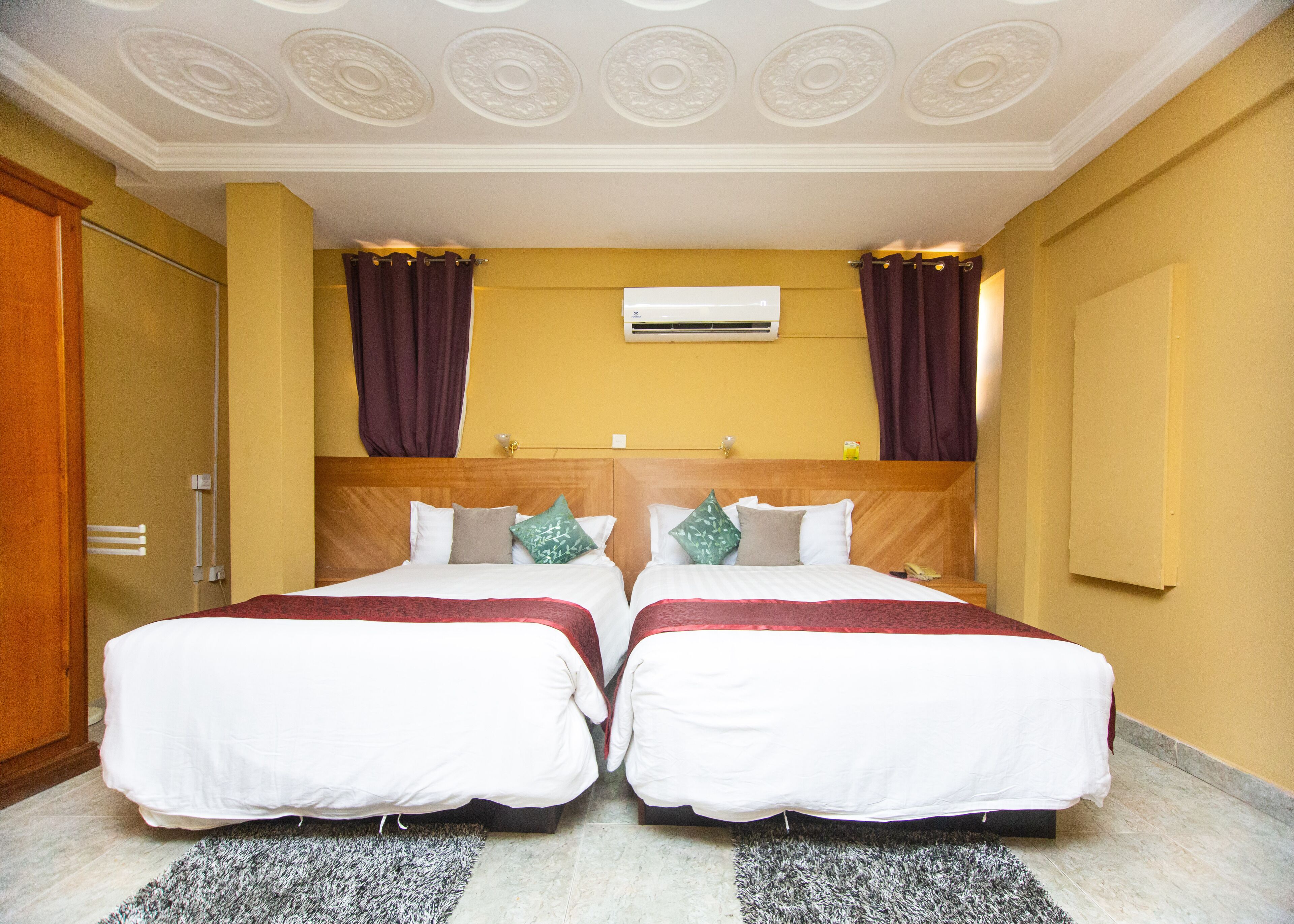 Habitación de hotel con dos camas, cabecera de madera, techo con molduras decorativas y aire acondicionado de pared.