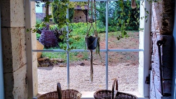 A rustic interior with a large window overlooking a garden, two wicker baskets on the floor, and hanging planters.