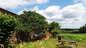 A rustic outdoor setting with a wooden table and chairs, a stone wall, and a large tree providing shade.