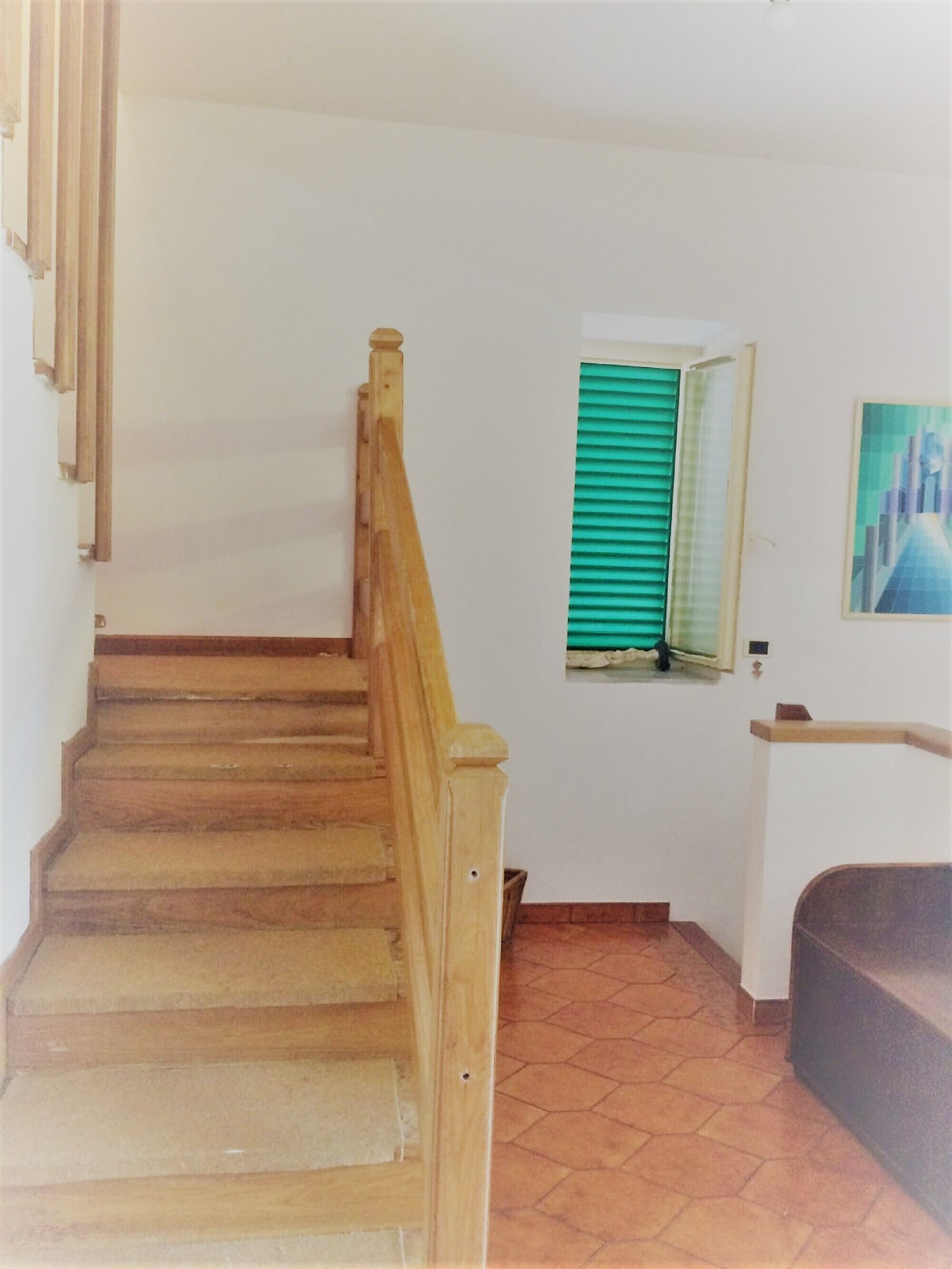 A wooden staircase with a handrail, a window with green shutters, and a painting on the wall.