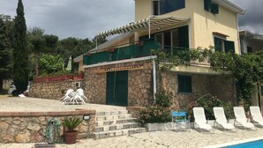 A two-story house with a stone facade, a balcony with green shutters, and a pool area with lounge chairs.