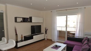 Una sala de estar moderna con televisor de pantalla plana, sofás blancos, una mesa de centro y un balcón con vistas.