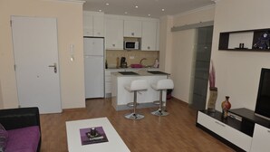 Un espacio habitable compacto con kitchenette, una zona de comedor con dos taburetes altos y una pequeña mesa blanca, además de un sofá púrpura.