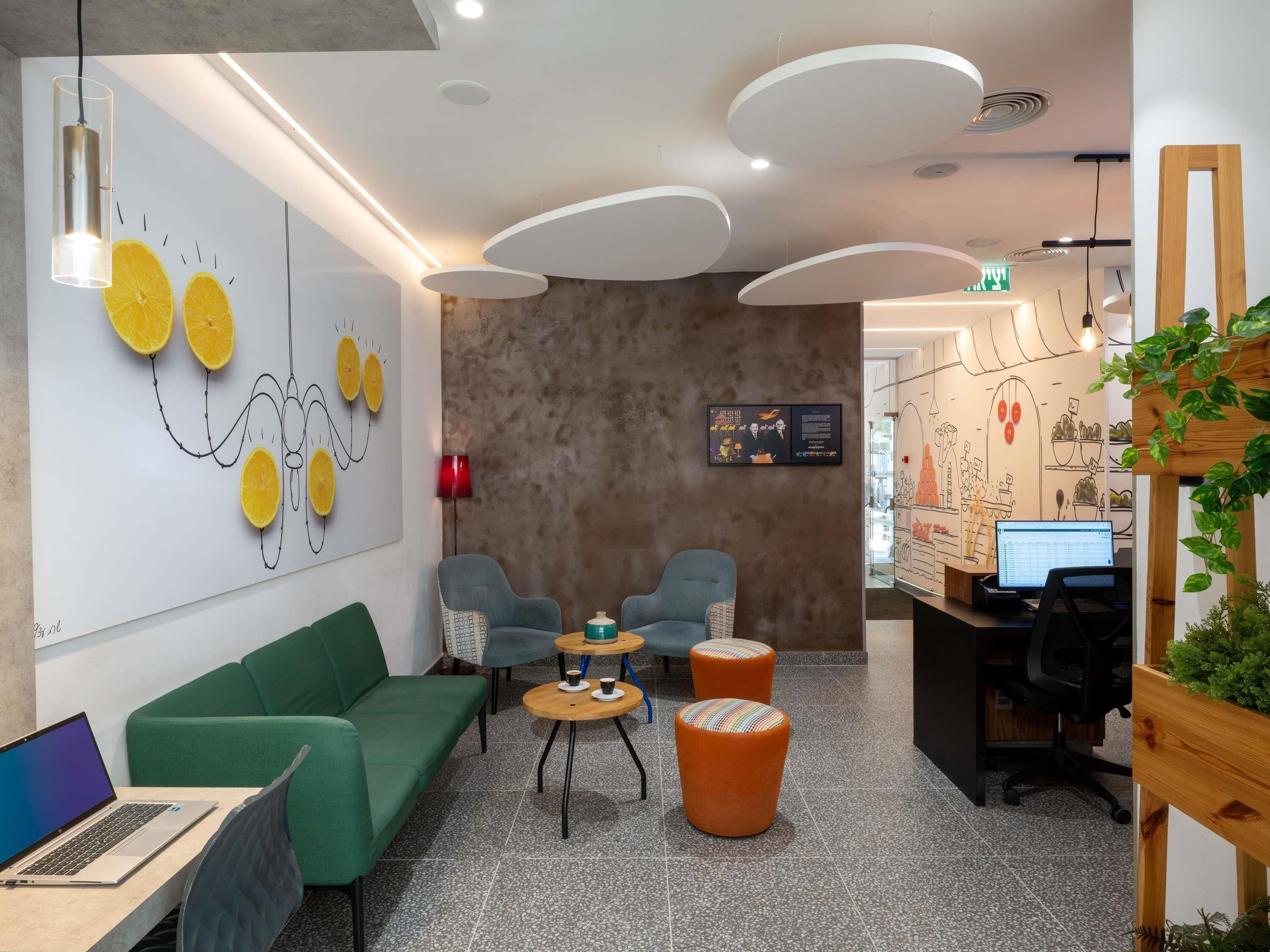 A modern hotel lobby with a green sofa, two armchairs, a coffee table, and a wooden desk with a computer.