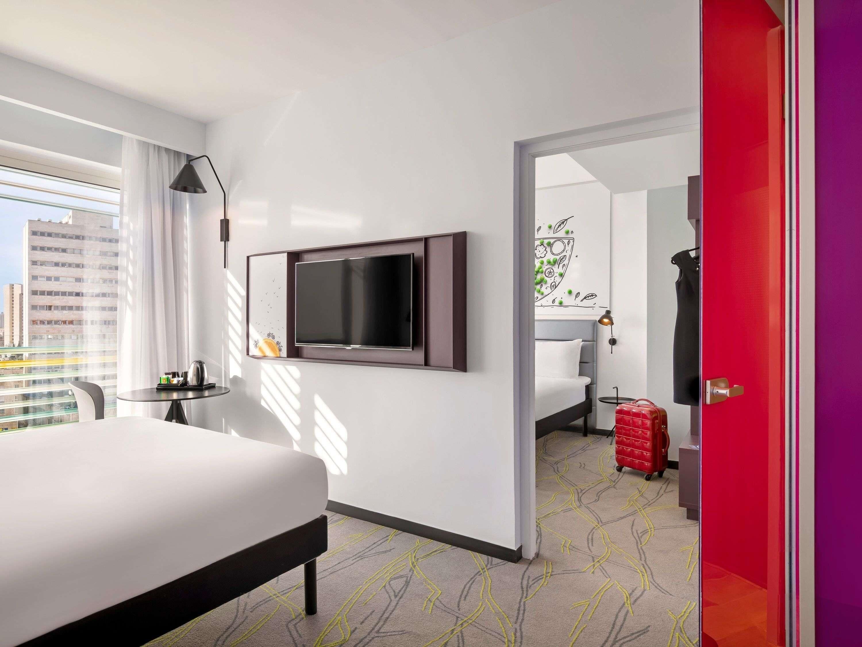 A hotel room with a bed, a TV mounted on the wall, a small table with a lamp, and a view of the city through the window.