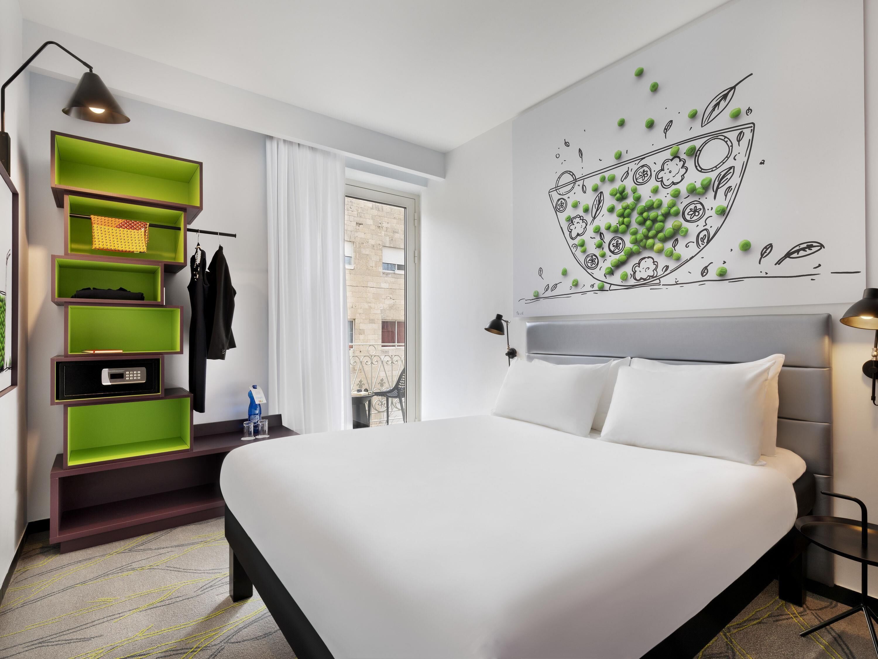 Una camera d'albergo moderna con un letto, una scaffalatura verde e un affresco al muro.