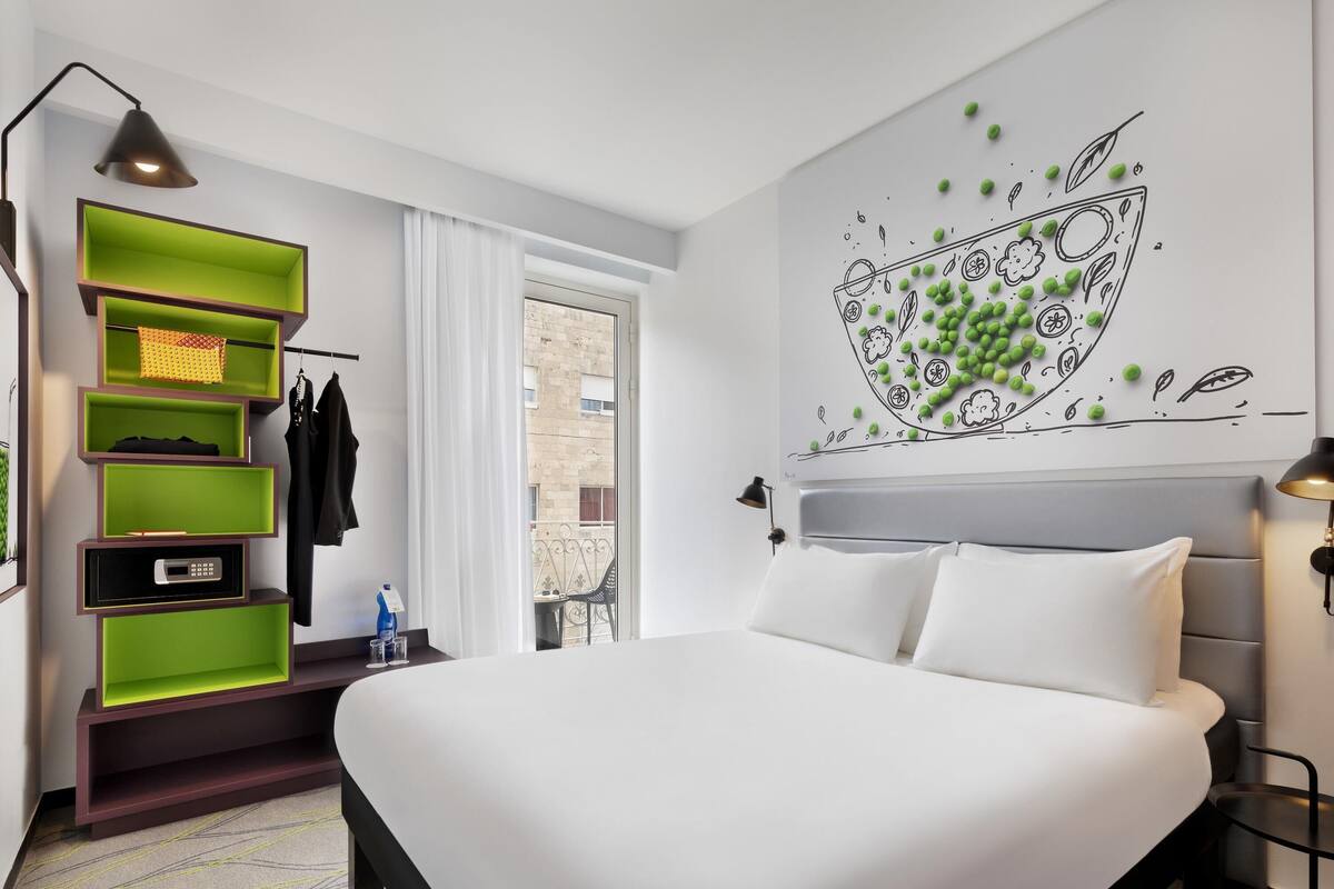 A modern hotel room with a bed, green shelving unit, and a wall mural.