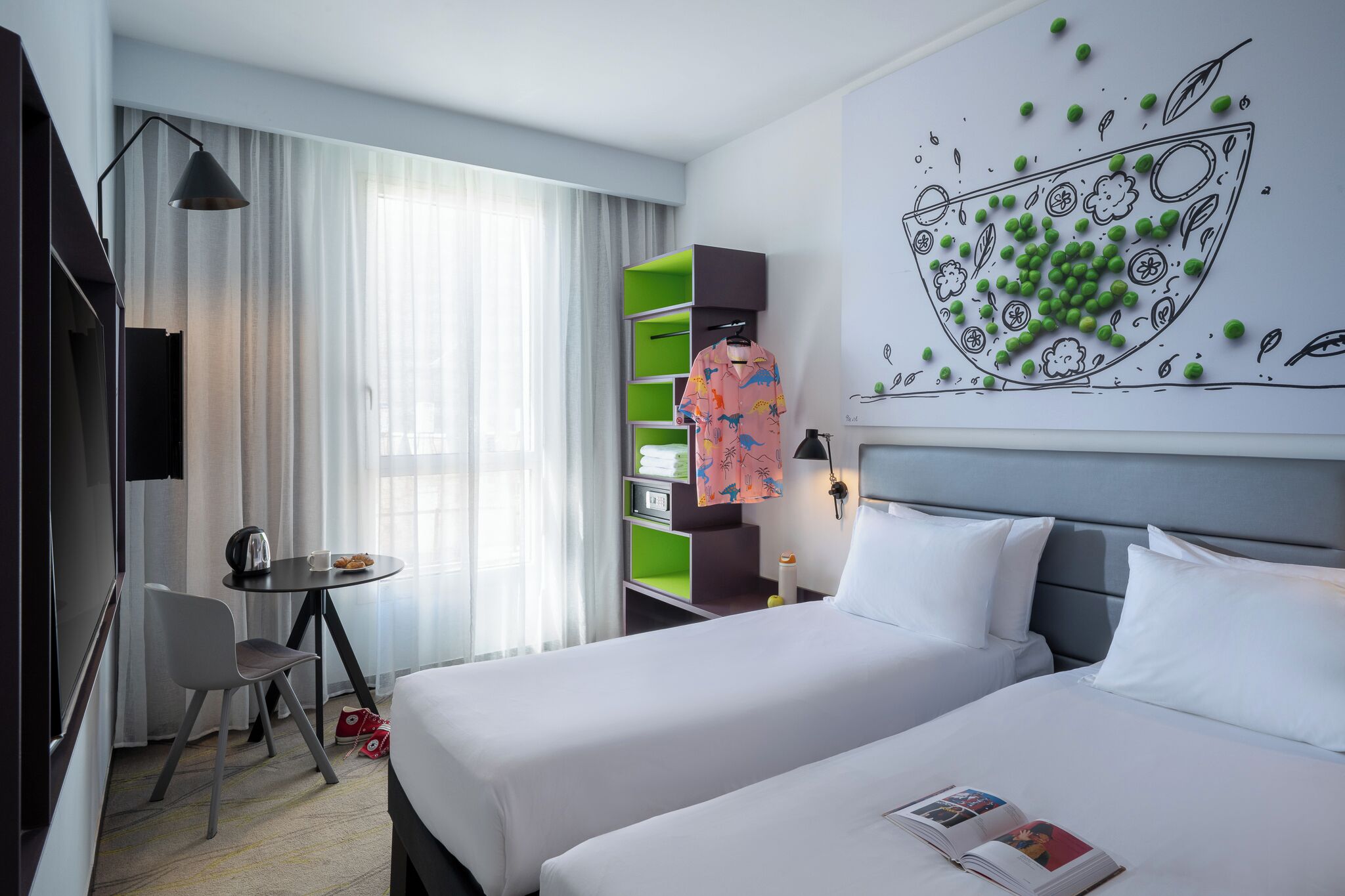Une chambre d’hôtel avec deux lits, un bureau avec une chaise, une petite table avec une tasse et des pâtisseries, et une étagère verte.