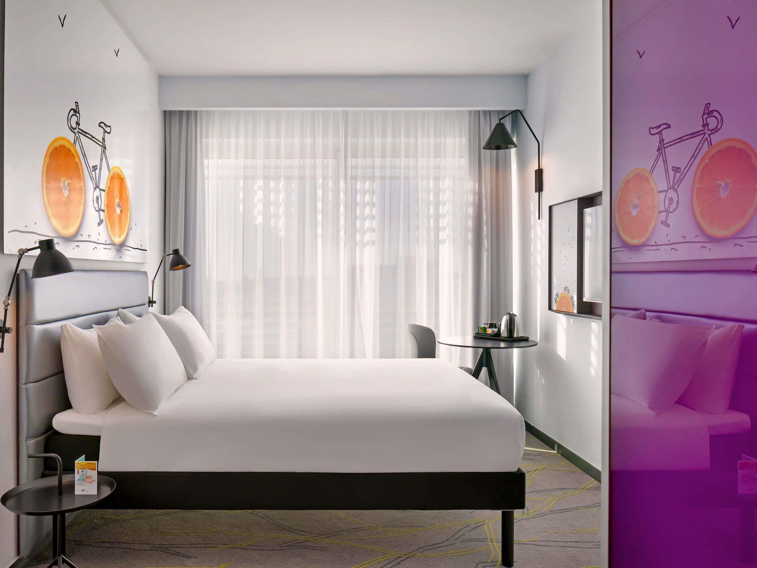 Habitación de hotel moderna con una cama grande, una mesita y una silla. Hay dos lámparas de pared y una ventana con cortinas traslúcidas.