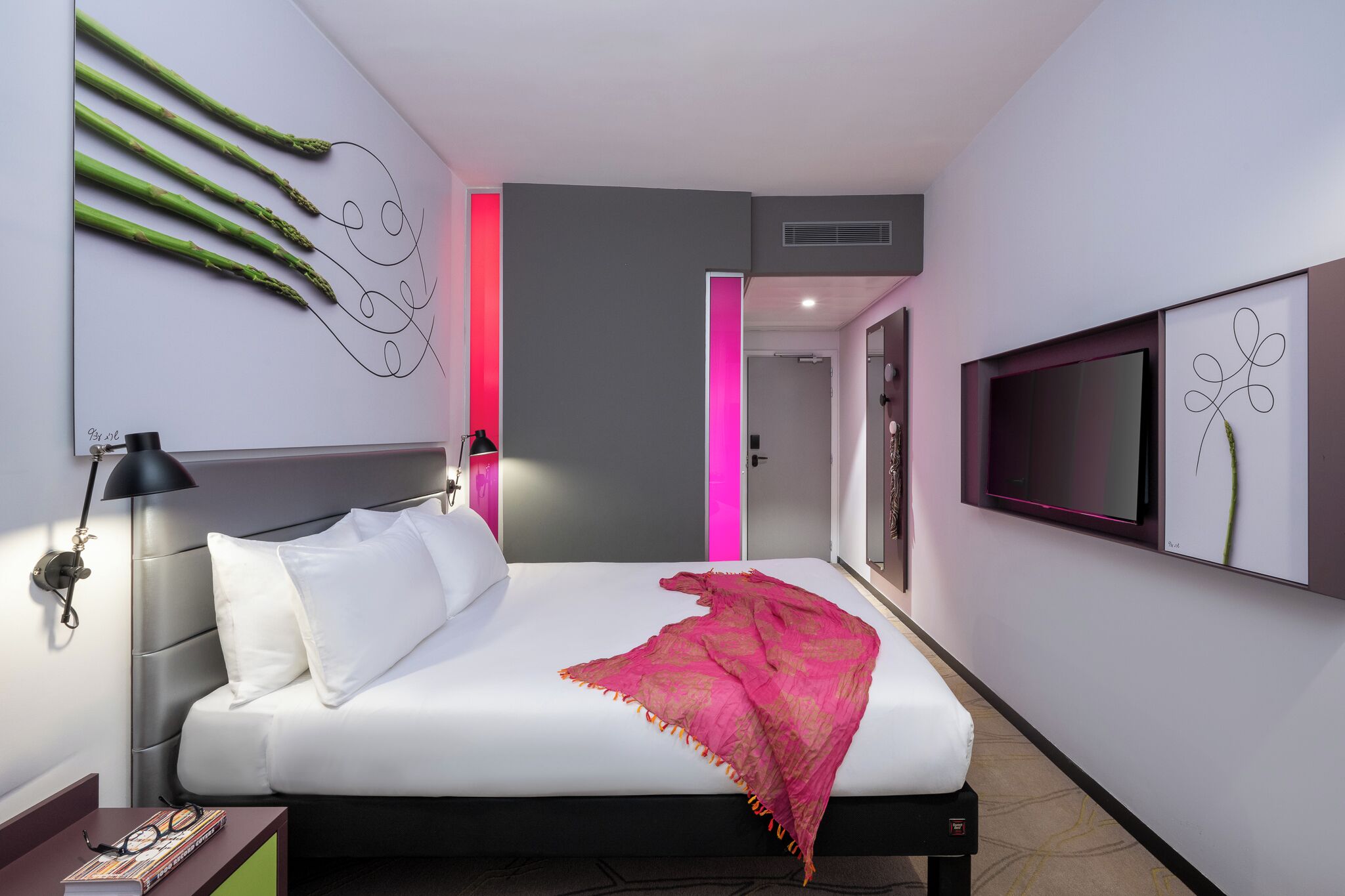 A modern hotel room with a bed, a desk, a TV, and a wall-mounted artwork.