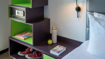 A hotel room with a bed, a nightstand with a lamp, a green shelf with a phone and a book, a shoe rack, and a water bottle.