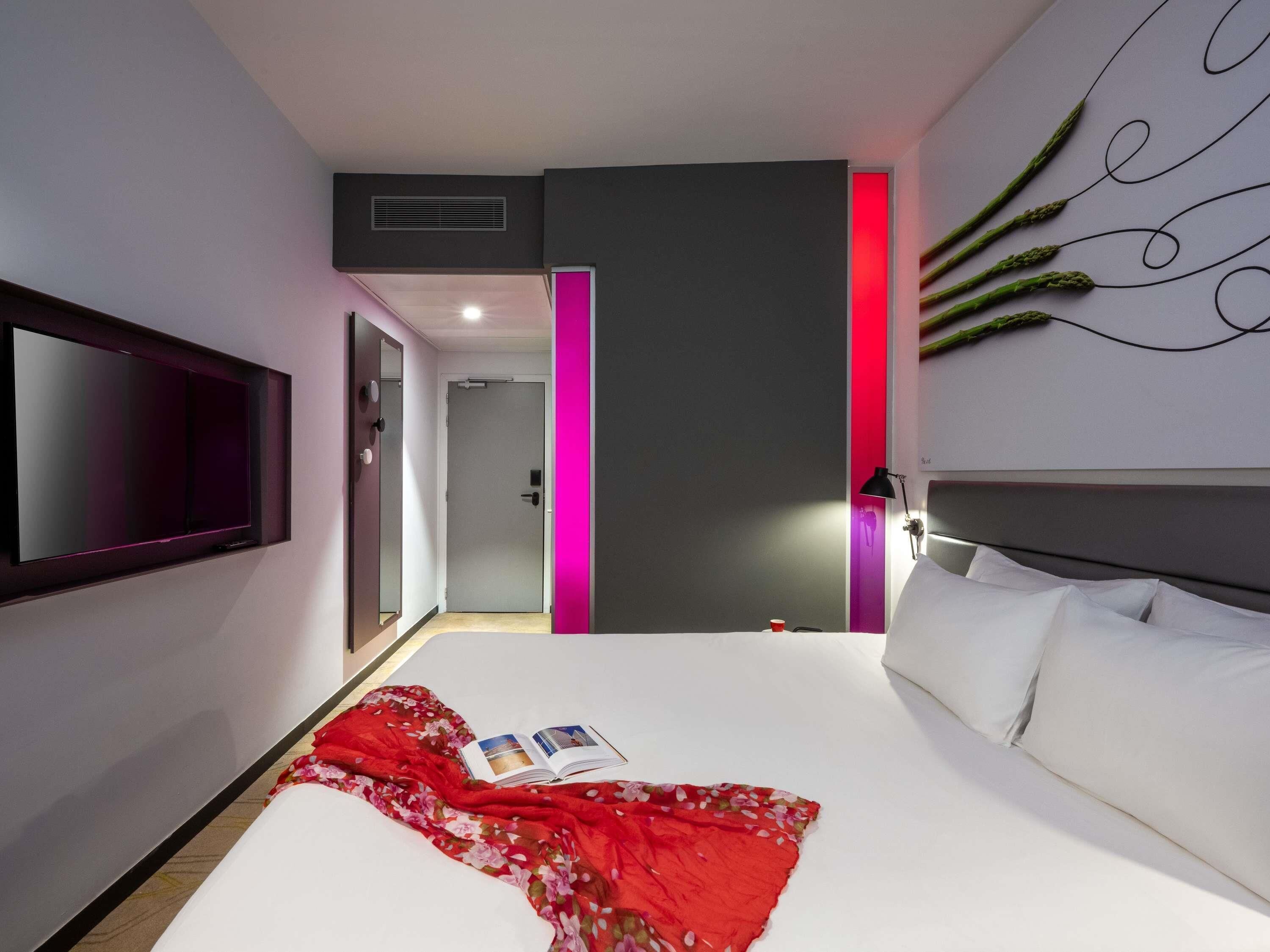 A modern hotel room with a large bed, a flat-screen TV mounted on the wall, and a bedside table with a lamp.