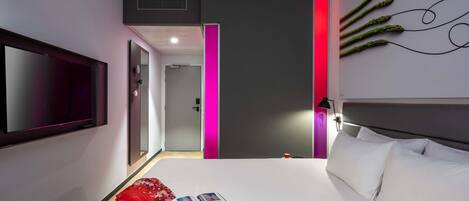 A modern hotel room with a large bed, a flat-screen TV mounted on the wall, and a bedside table with a lamp.