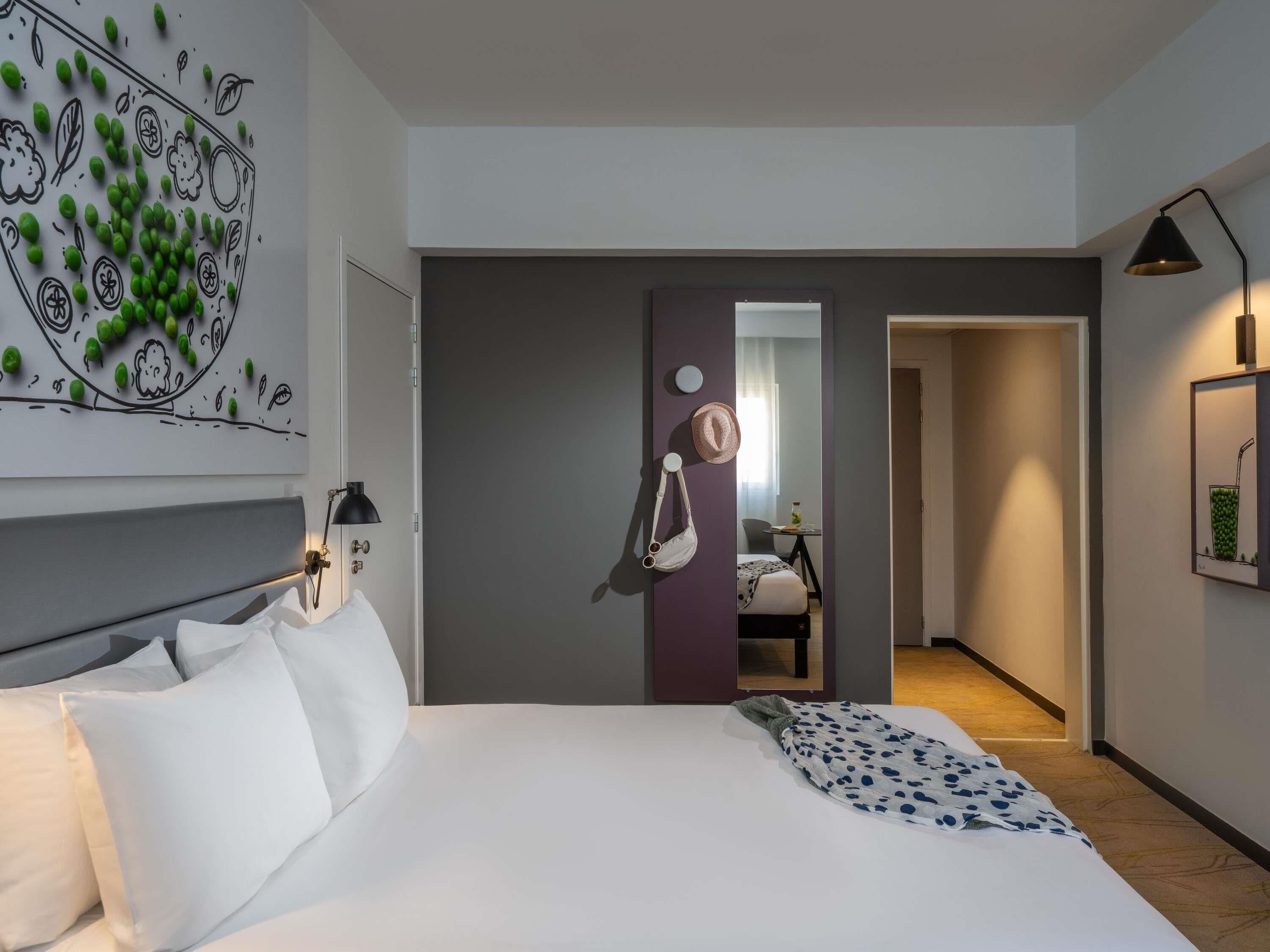Une chambre d’hôtel moderne avec un grand lit, une table de chevet, une œuvre d’art murale, un miroir et un couloir menant à une autre chambre.