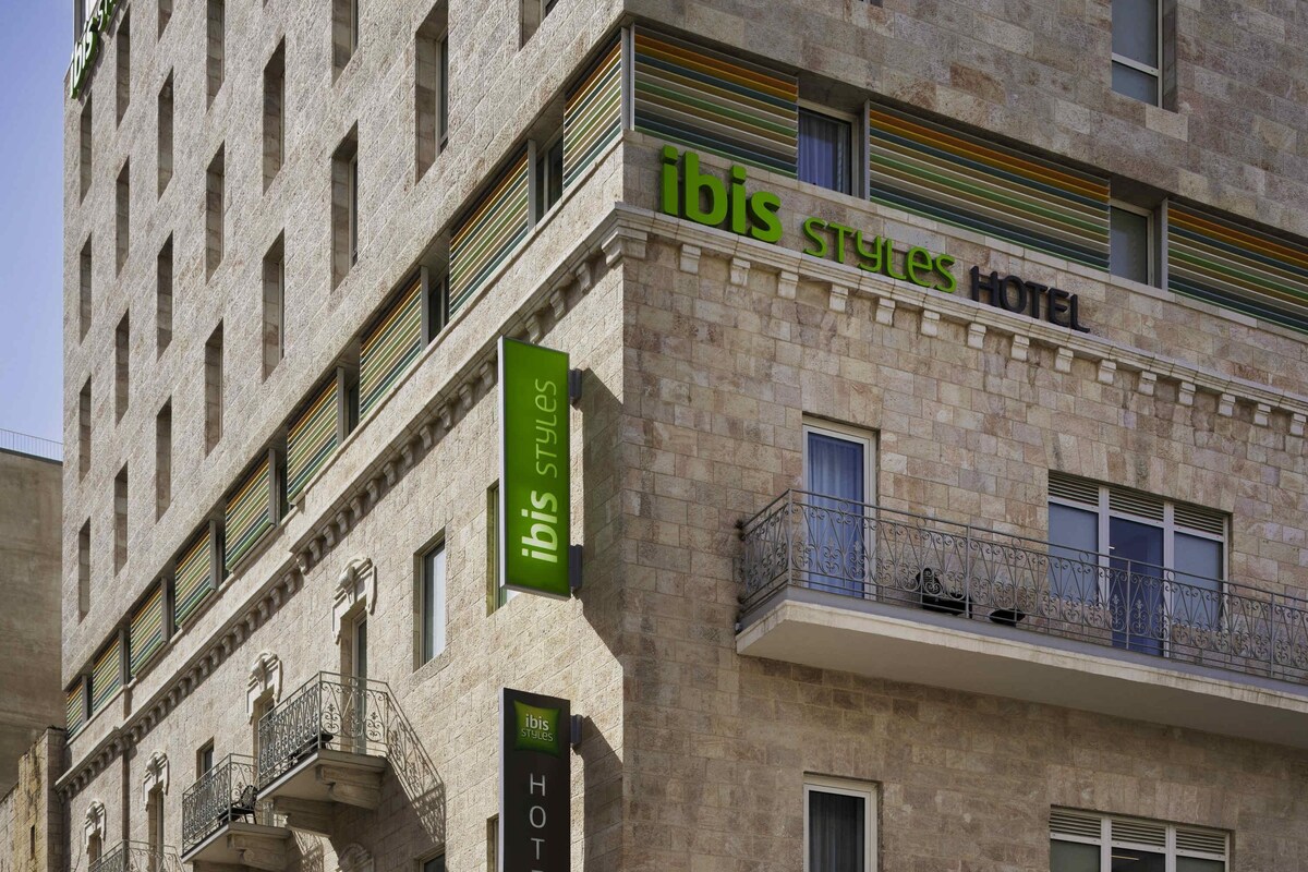 Ein Hotel mit Steinfassade, mehreren Balkonen mit schmiedeeisernen Geländern und Beschilderung, die es als Teil der ibis Styles Hotelkette kennzeichnet.