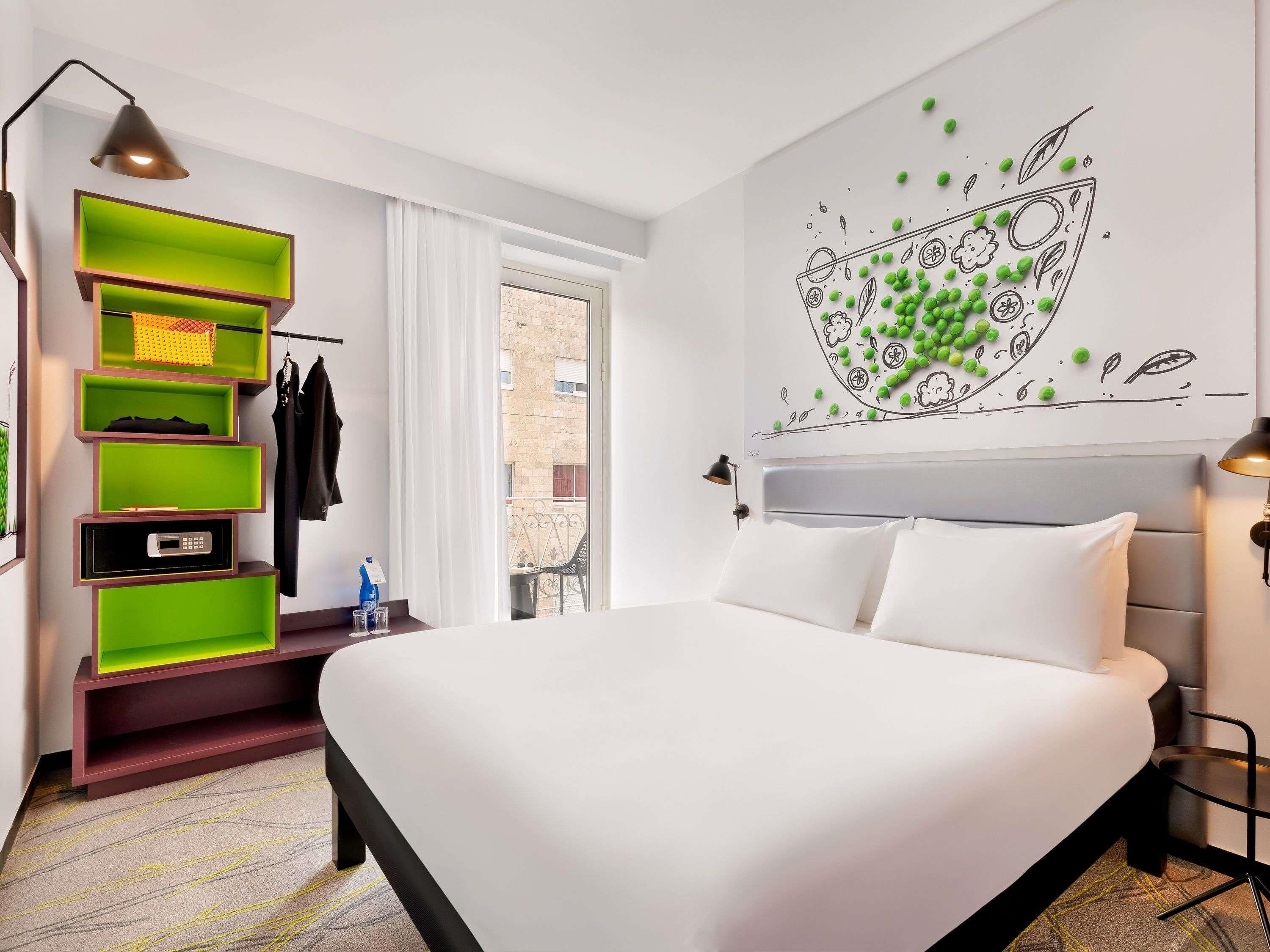 A modern hotel room with a bed, a wall-mounted shelf unit, a window with a view, and a wall art piece.