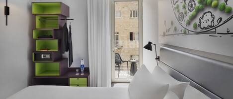 A hotel room with a bed, a desk with a computer, a green storage unit, a window with a view of the outside, and a wall with a green design.