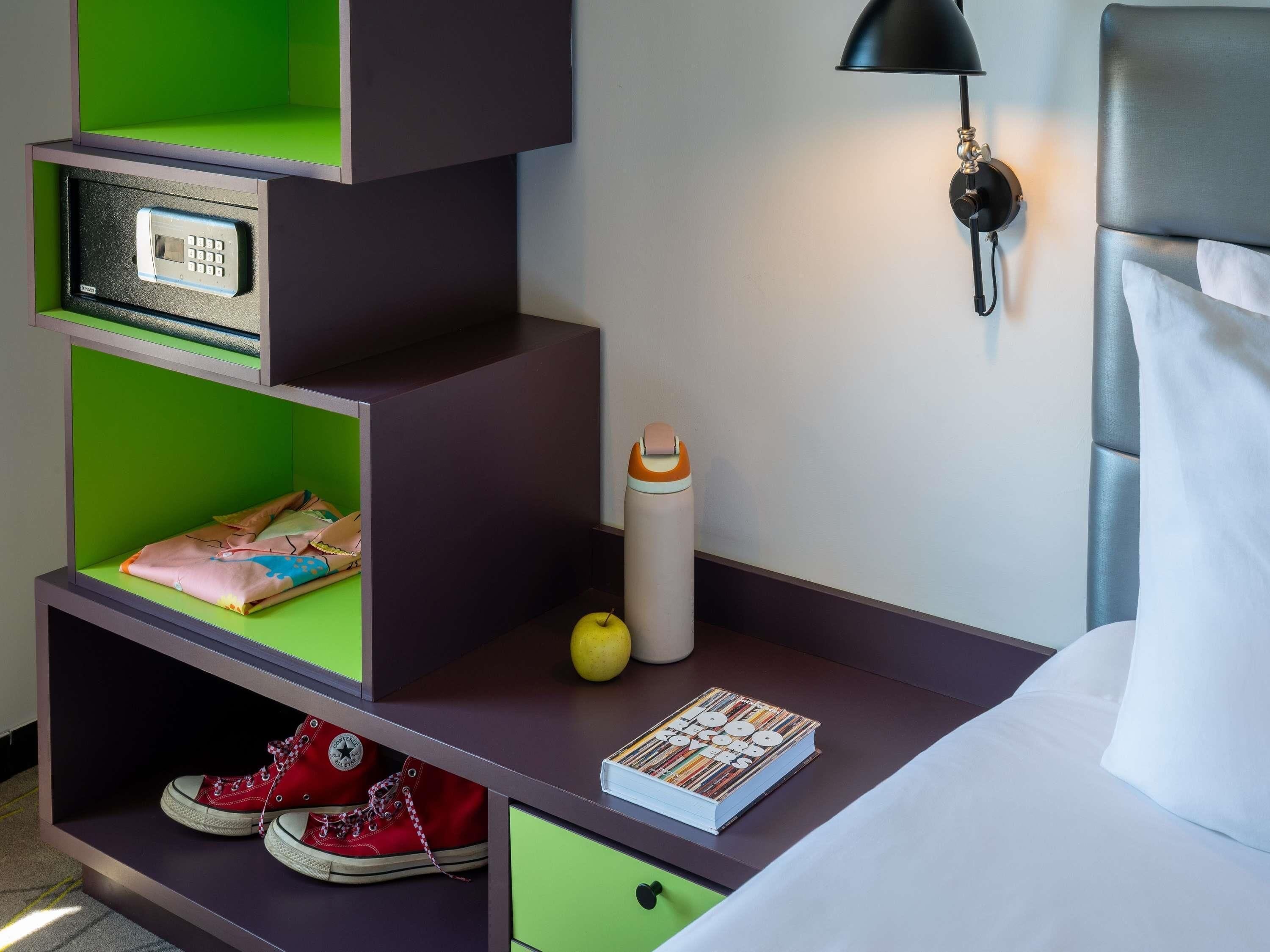 A hotel room with a bed, a nightstand with a lamp, a green storage unit with shoes and a book, and a wall-mounted phone.