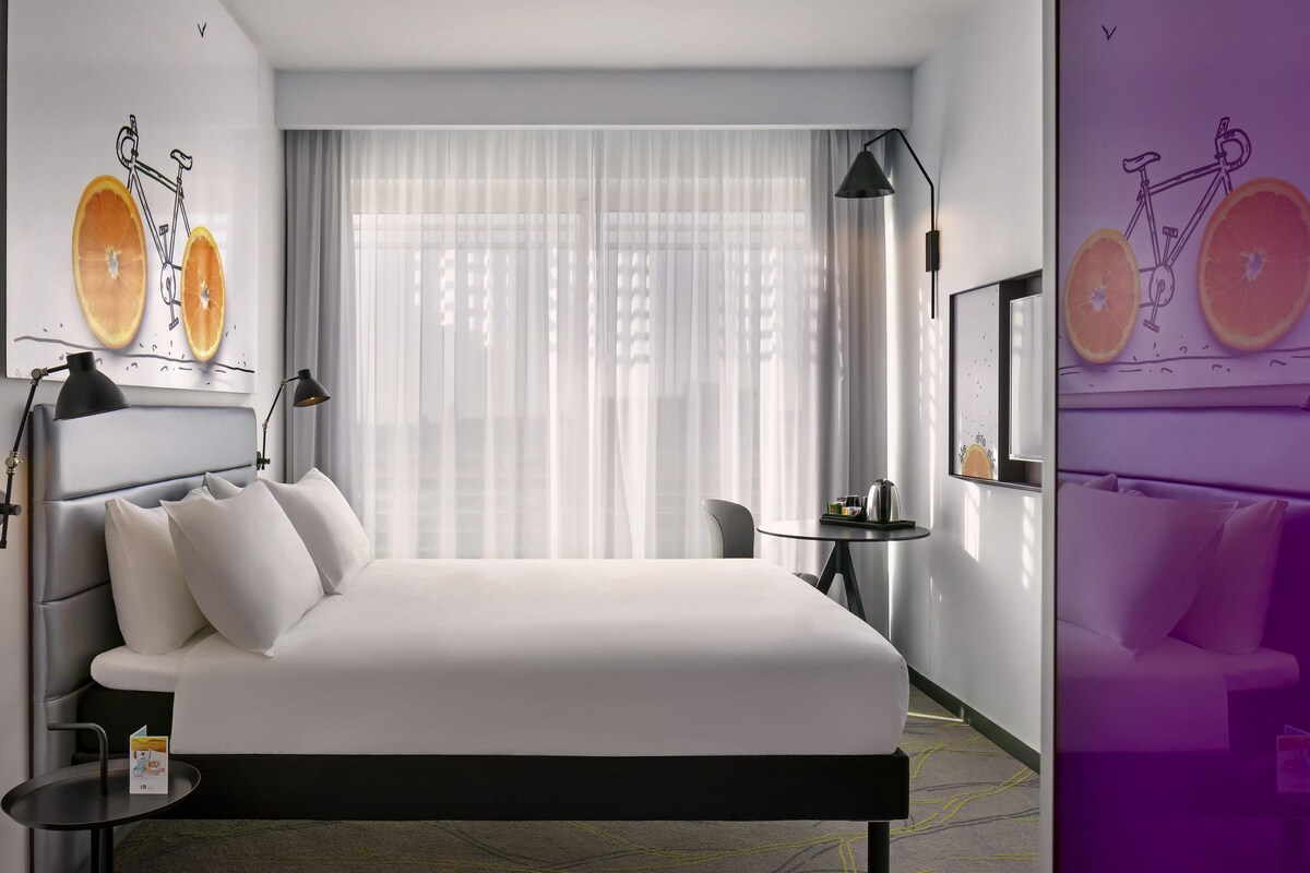 A modern hotel room with a large bed, a small table, a chair, and a window with sheer curtains.