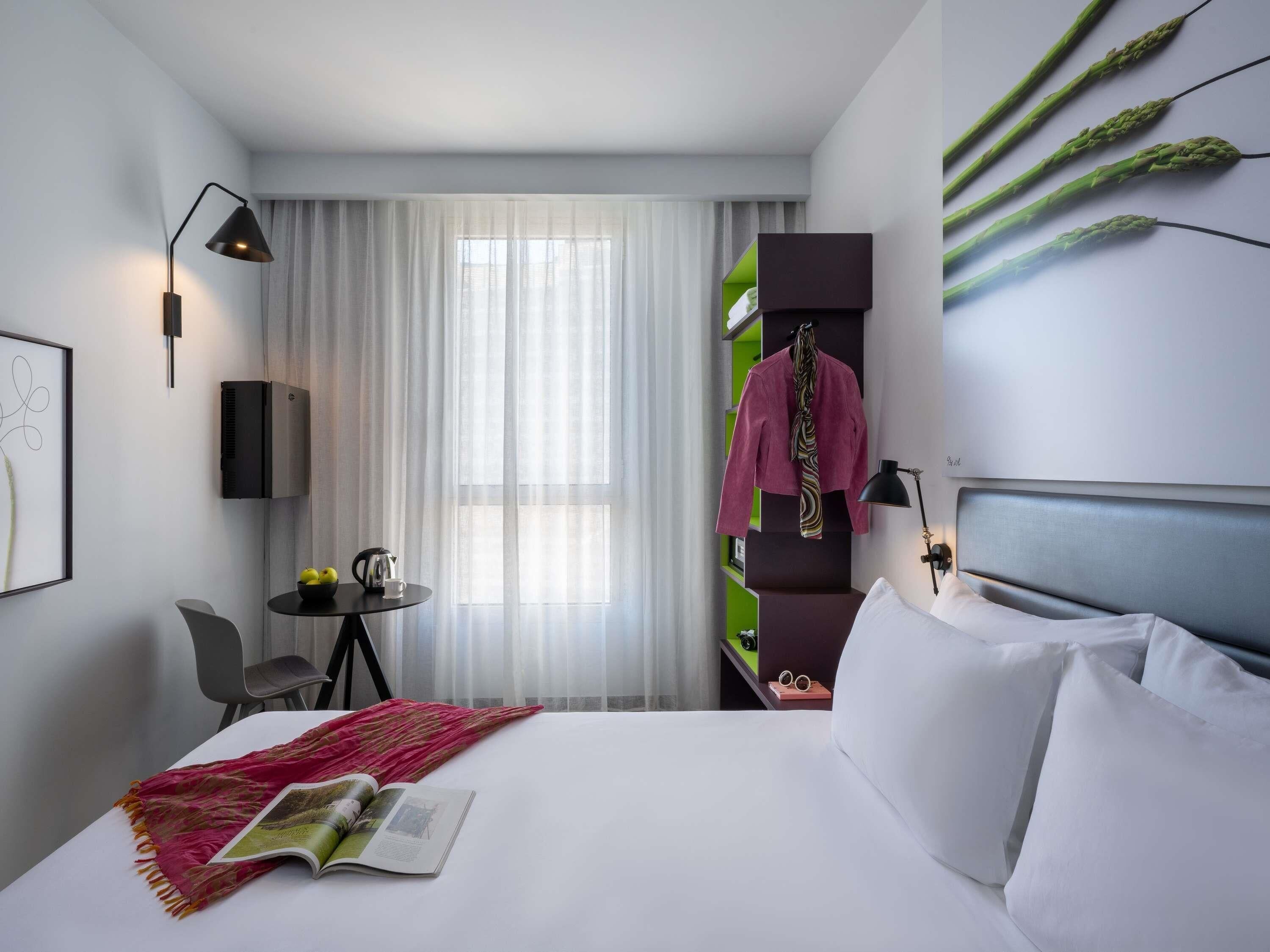 A hotel room with a bed, a reading area with a magazine, a table with a kettle, and a window with curtains.