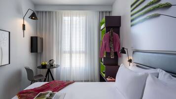 A hotel room with a bed, a reading area with a magazine, a table with a kettle, and a window with curtains.