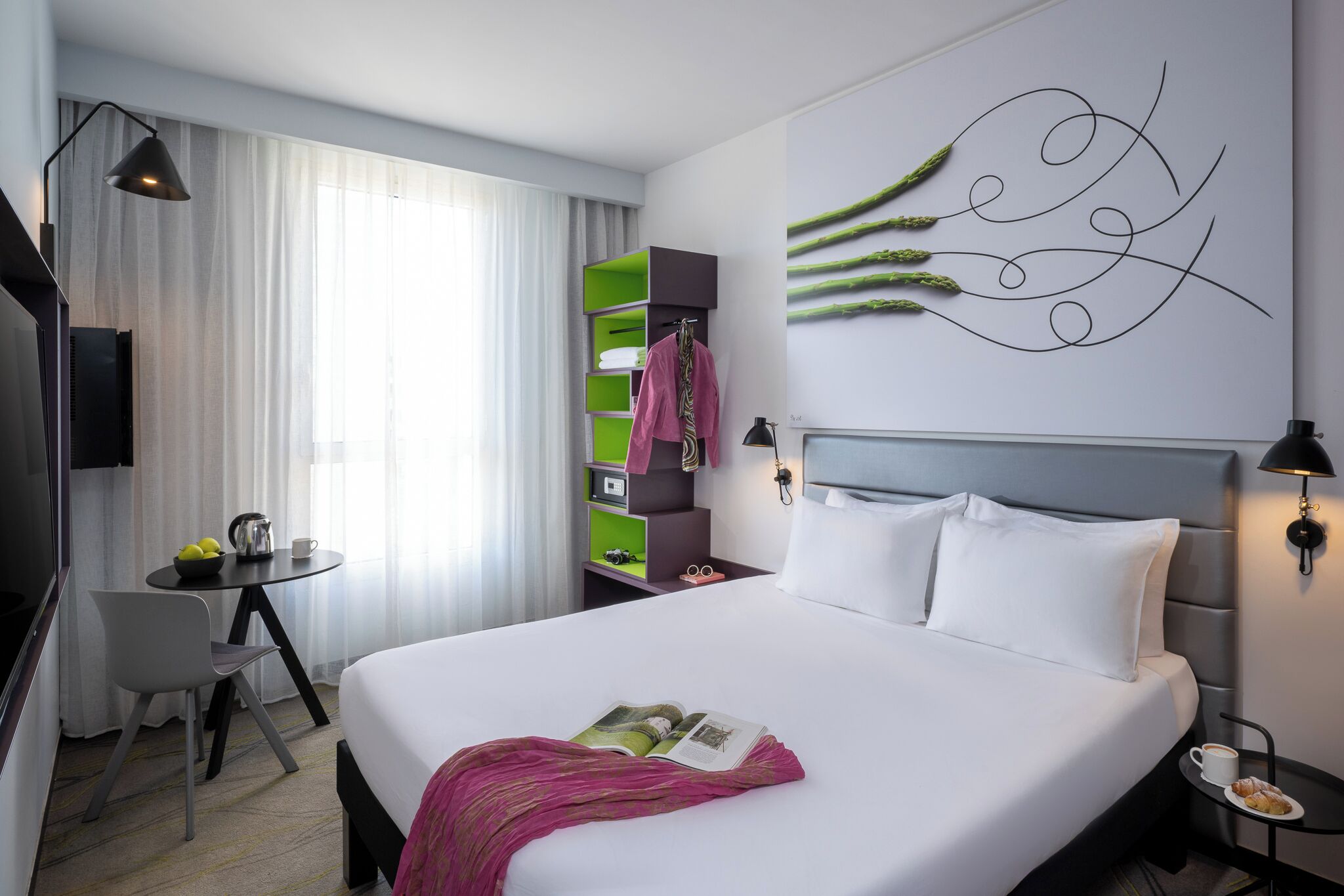 A hotel room with a bed, a desk, a chair, a small table with a cup and a bowl of fruit, a wall-mounted lamp, and a decorative wall piece.