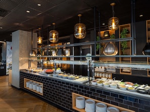 A buffet area with various food options, a counter with plates, and hanging pendant lights. - ibis Styles Jerusalem City Center - An AccorHotels Brand (Jerusalem)