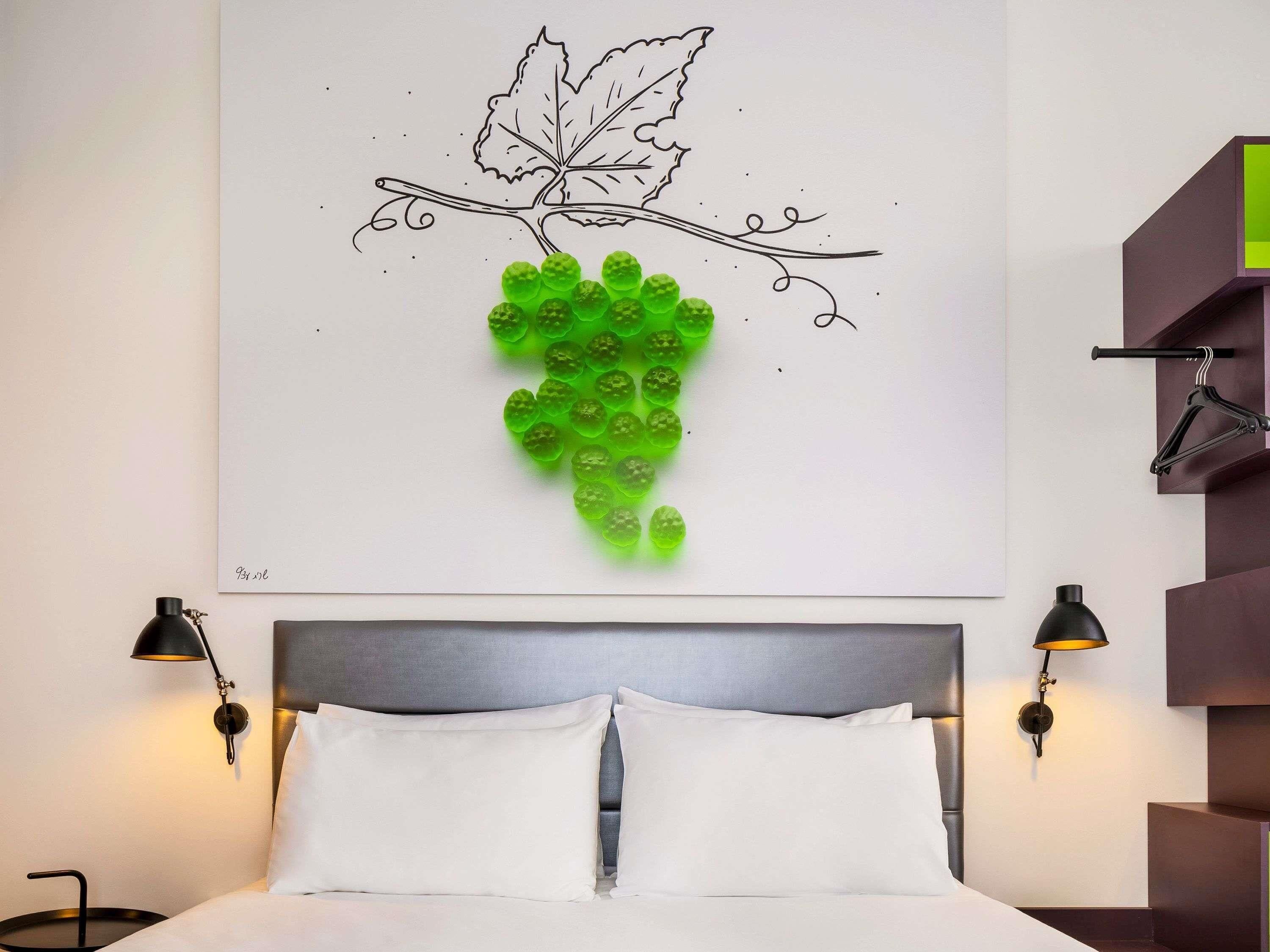 A bedroom with a bed, two bedside lamps, a wall art of a grapevine, and a hanging coat rack.