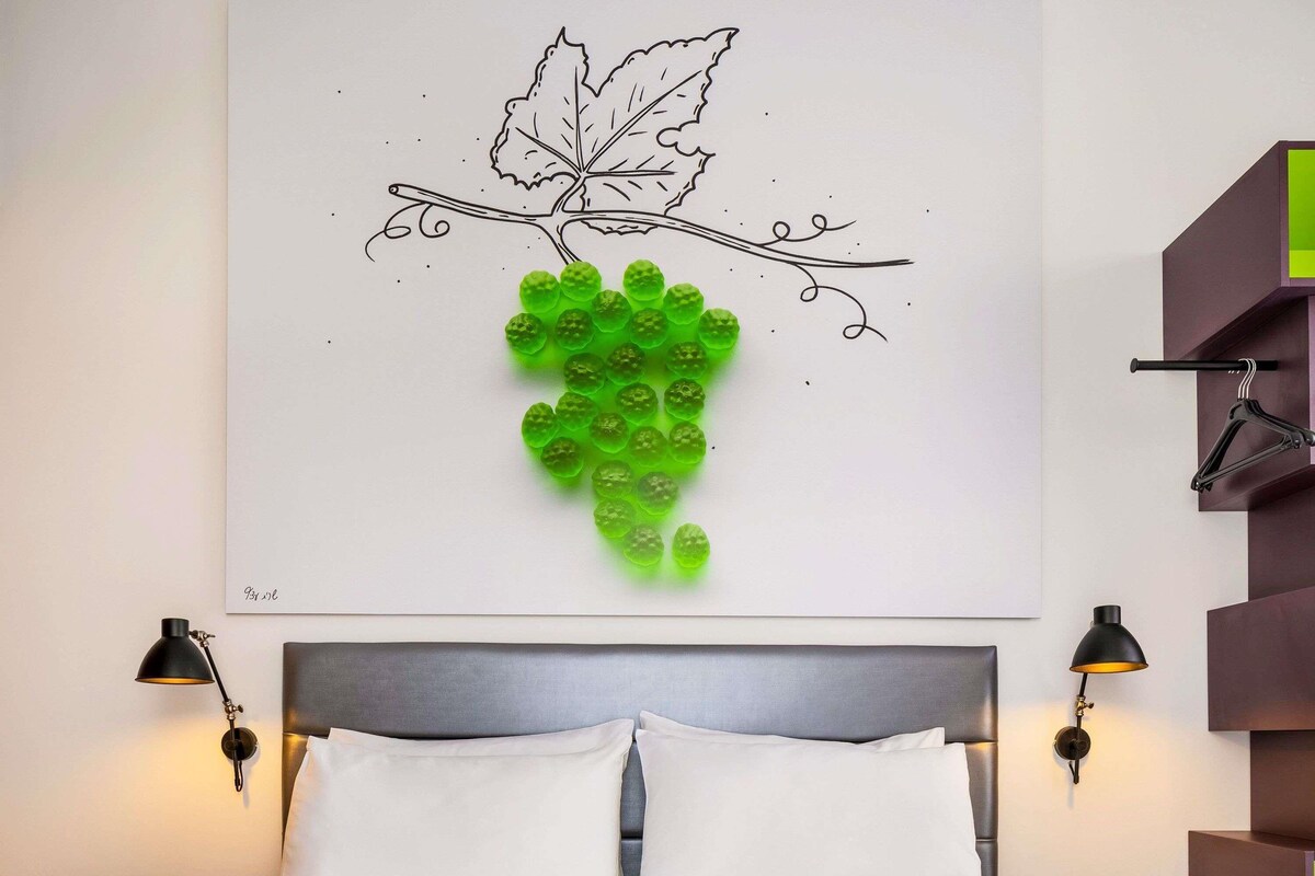 Ein Schlafzimmer mit einem Bett, zwei Nachttischlampen, Wandbild mit einem Weinstock und einem hängenden Kleiderständer.